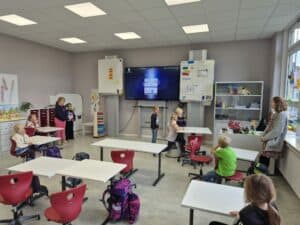 Bunte Klassenraumansicht der GS Mühlenbergschule Osloß mit Kindern, Lehrerin und moderner digitaler Ausstattung, ideal für multikulturelles Lernen und innovative Bildungsangebote.